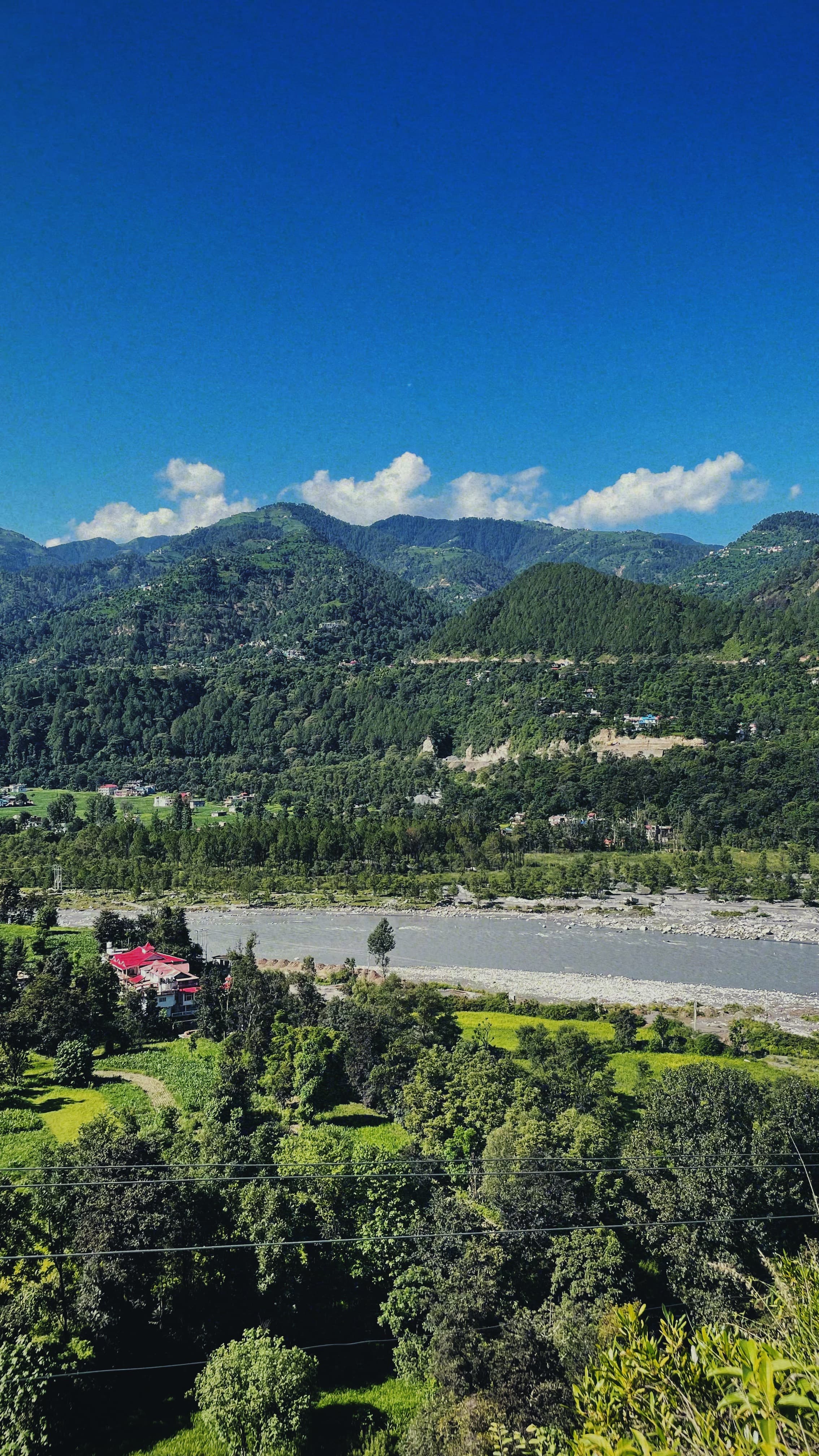A breathtaking aerial view of Jimmy's River Resort Chamba, showcasing its central building amidst green fields, with the River Ravi flowing through a tranquil valley surrounded by wooded hills.