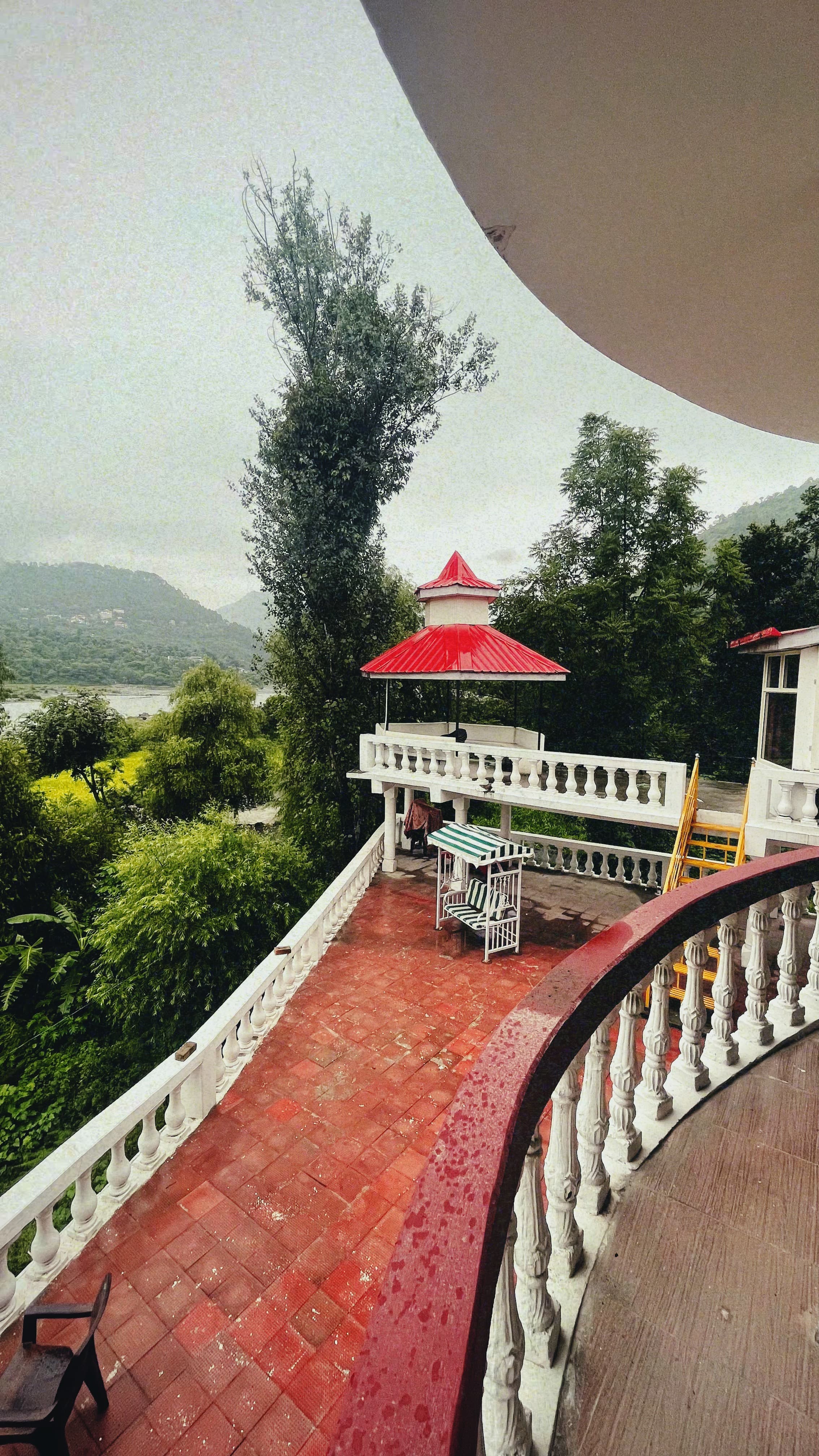 Private balcony at Jimmy's River Resort Chamba, featuring a cozy outdoor seating area with white railings, overlooking lush greenery and hills under an overcast sky.