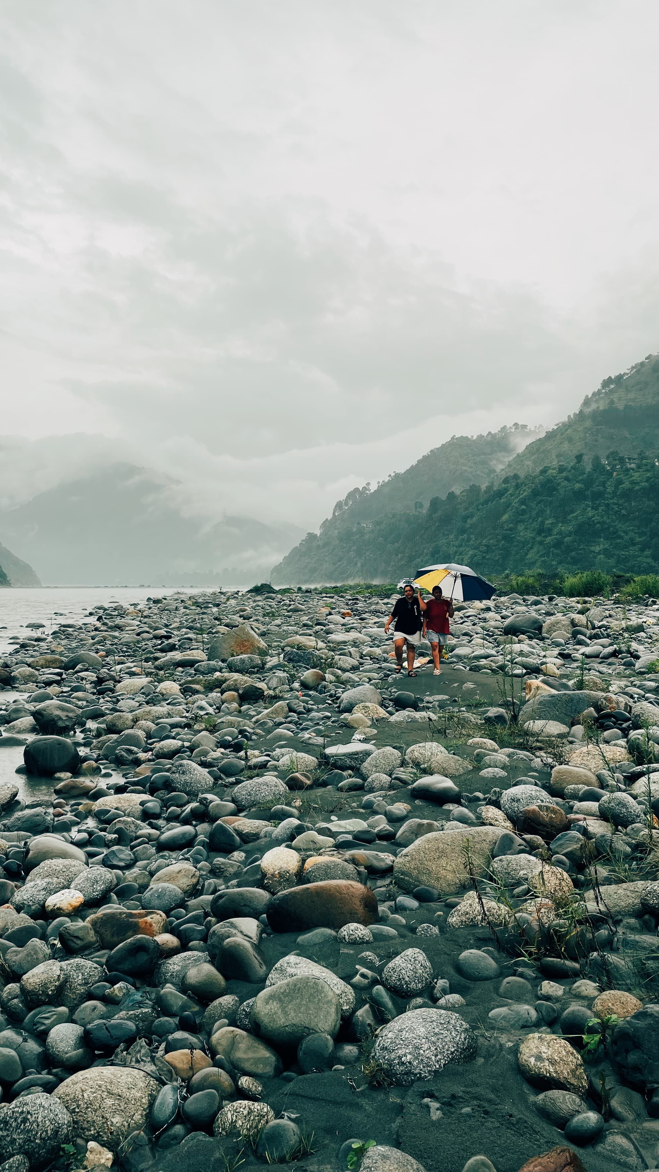 Scenic riverside at Jimmy's River Resort Chamba, with a couple walking under an umbrella along the smooth stones of the River Ravi, framed by misty hills and lush vegetation.