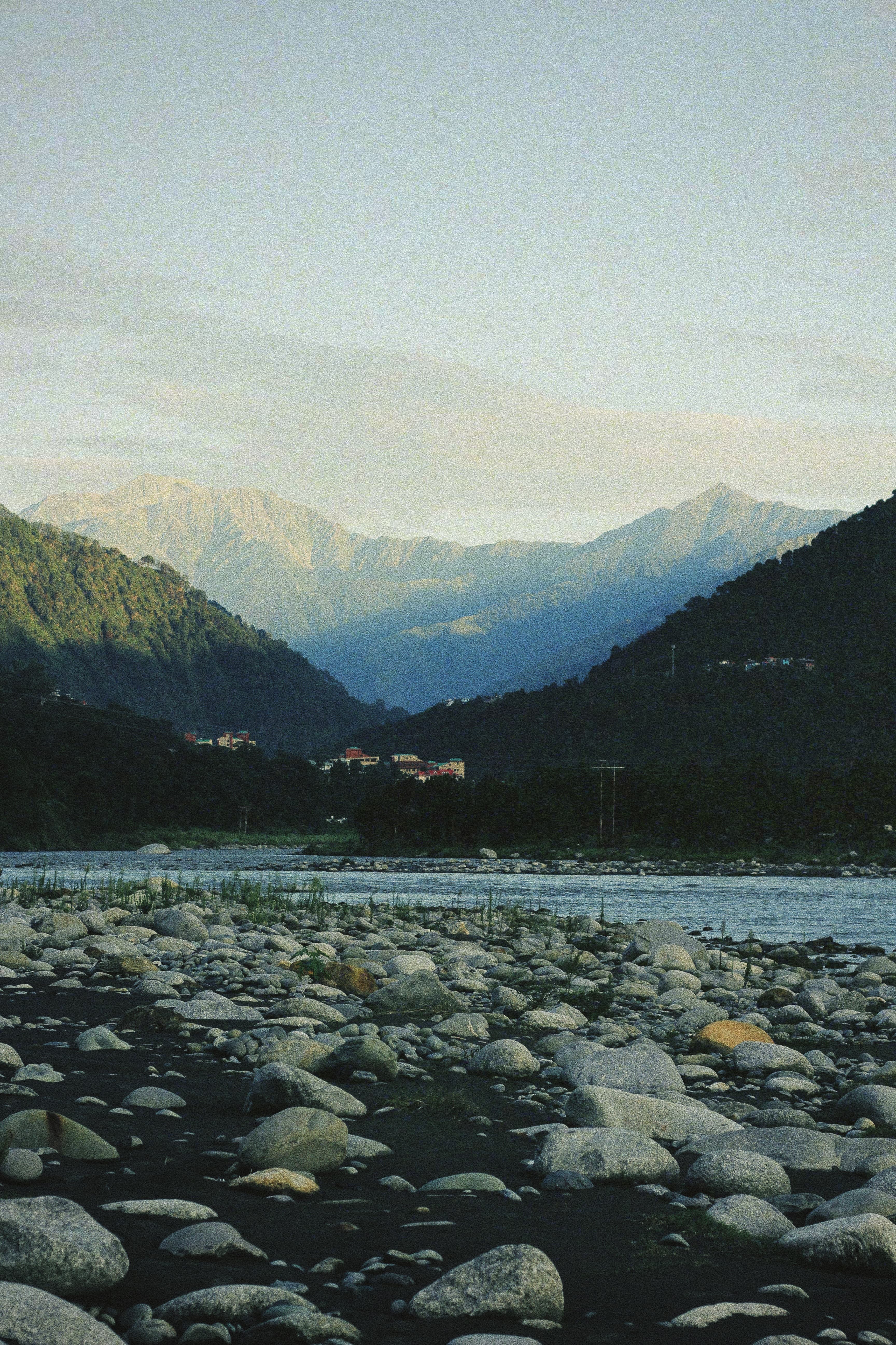 Picturesque view of the River Ravi at Jimmy's River Resort Chamba, with smooth stones scattered along the riverbank, lush green hills framing the scene, and distant mountains under a tranquil sky.