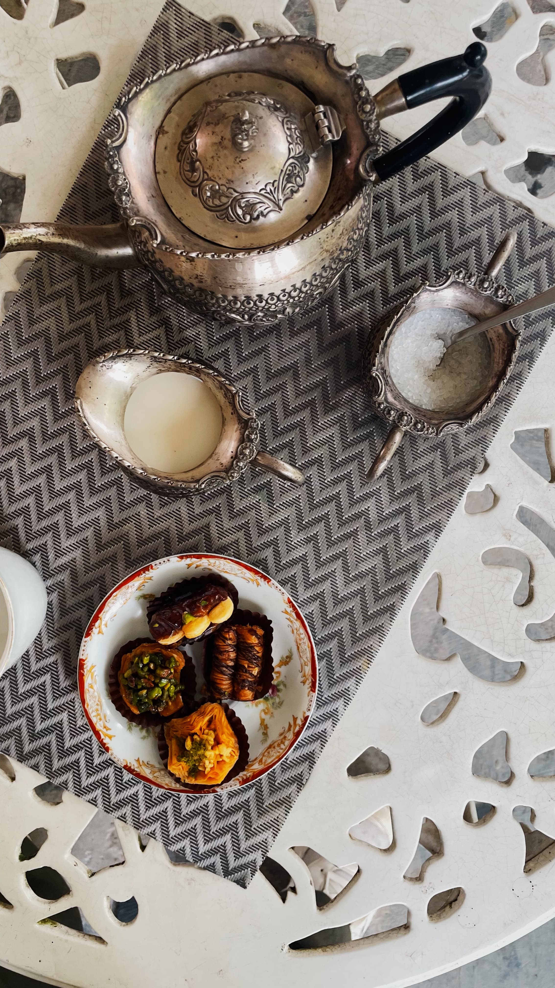 Elegant tea setup at Jimmy's River Resort Chamba, showcasing ornate silver teaware on a patterned fabric mat and a plate of assorted Middle Eastern sweets, placed on a decorative white table.